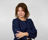 a woman in a dark dress with her arms folded, against a light, plain background