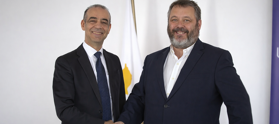 Two men in suits with the Cypriot and Polish flags in the background