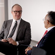 Two men in suits talking; the man in the foreground is seen from behind