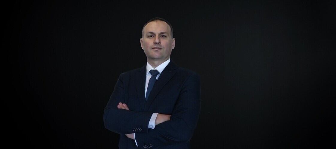 Męzczyzna w ciemnej marynarce, jasnej koszuli i ciemnym krawacie na ciemnym jednolitym tle