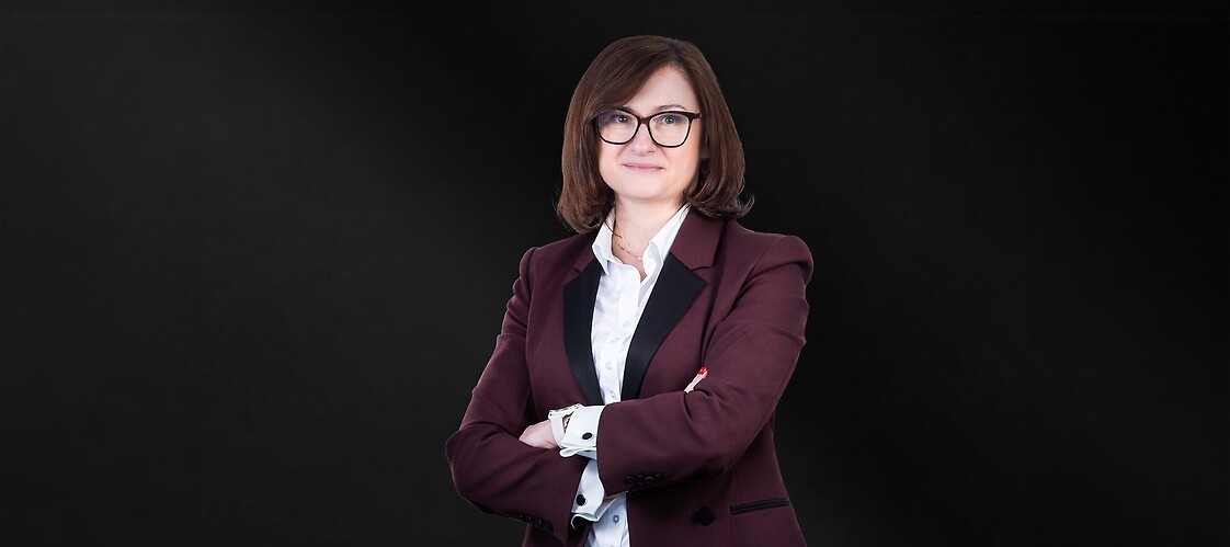 Kobieta w okularach, w ciemnobrązowym żakiecie i jasnej koszuli na ciemnym jednolitym tle