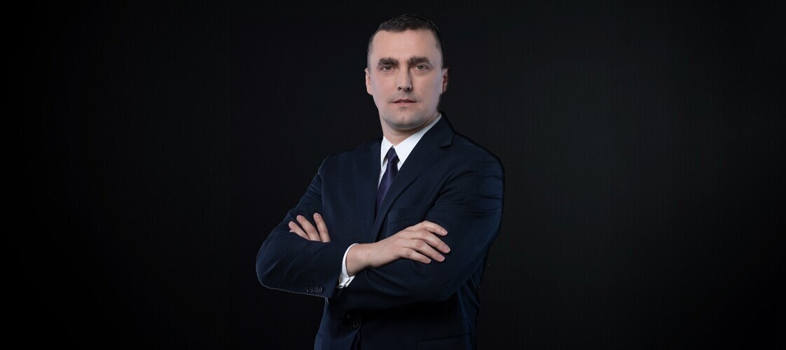 Mężczyzna w ciemnej marynarce, białej koszuli i xiemnym krawacie na ciemnym jednolitym tle