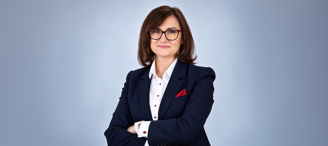 Kobieta w ciemnym żakiecie i jasne bluzce, z założonymi rękami, na jasnym tle