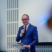 Mężczyzna w garniturze i białej koszuli, z mikrofonem w sali konferencyjnej