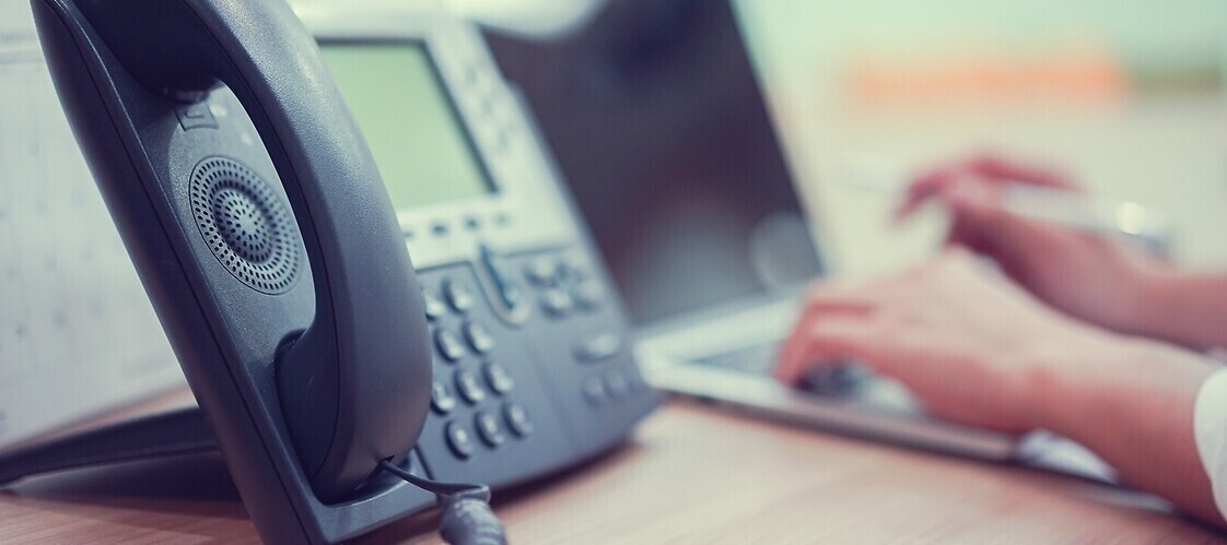 Telefon stacjonarny, w tle dłonie na klawiaturze kmpuera