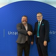 Dwóch mężczyzn w garniturach podaje sobie dłoń na tle bannera z logotypem UKE
