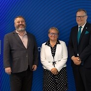 Dwóch mężczyzn oraz kobieta w w strojach biznesowych na tle bannera z logotypem UKE