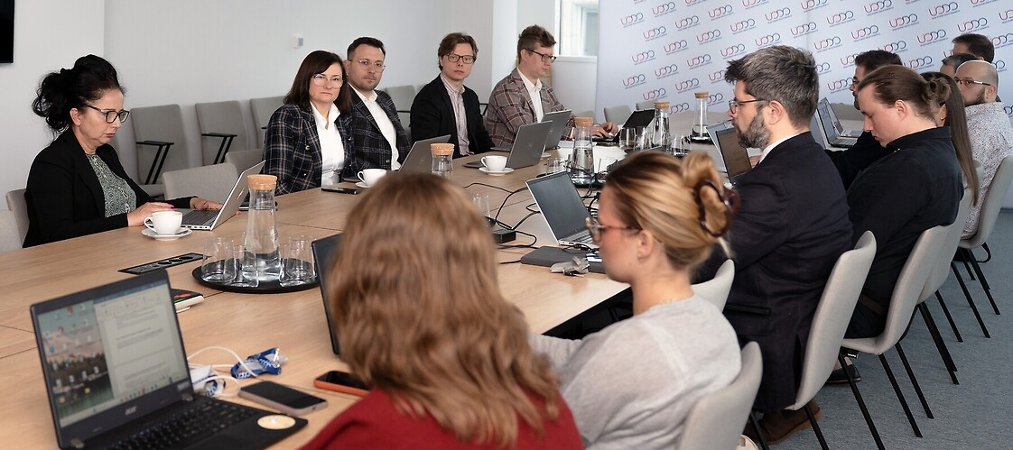 Grupa kilku osób w strojach biznjesowych po obu stronach stołu konferencyjnego