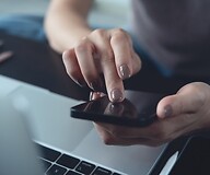 Dłoń na ekranie smartfona, poniżej otwarty laptop