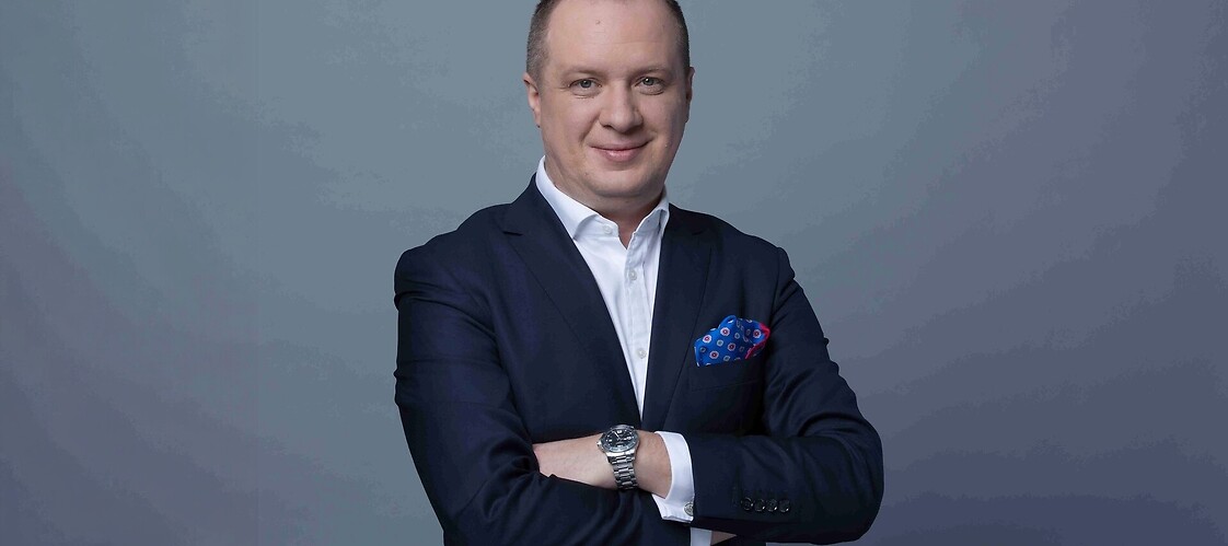 Mężczyzna w garniturze z założonymi rękami, na ciemnym jednolitym tle