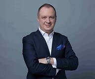Mężczyzna w garniturze z założonymi rękami, na ciemnym jednolitym tle