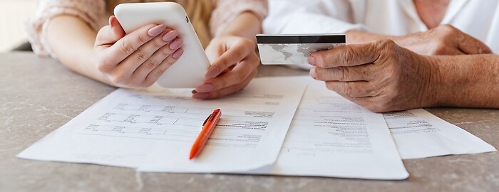 Dwie kobiety siedzą przed dokumentami. Jedna młodsza trzyma w ręku smartfona, ...