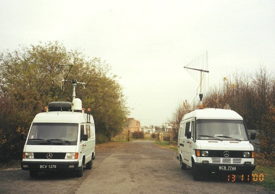 Ruchome stacje pomiarowe - dwa białe samochody (busy) z zamontowanymi antenami pomiarowymi na dachu.